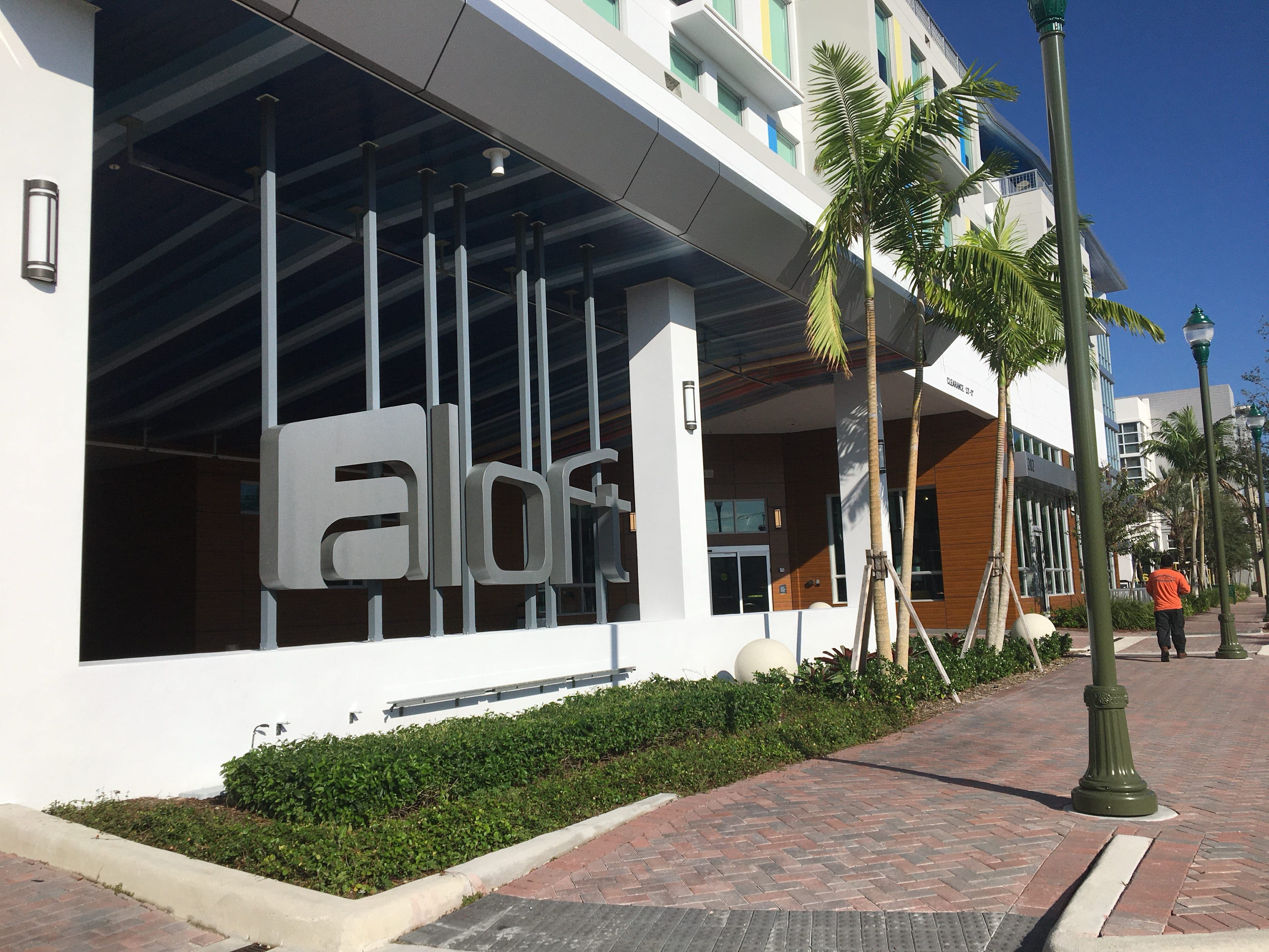 The Aloft hotel entrance canopy in Delray Beach, with sculptural signage, young royal palms in a planted bed, and a brick paver walkway