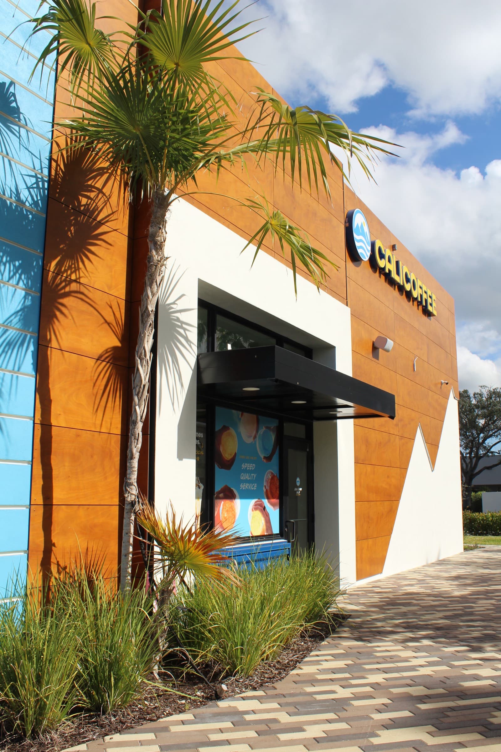 The Cali Coffee kiosk's wood-paneled fascia and covered order window under the CALI COFFEE sign, framed by a sabal palm and muhly grass along a patterned brick paver walk