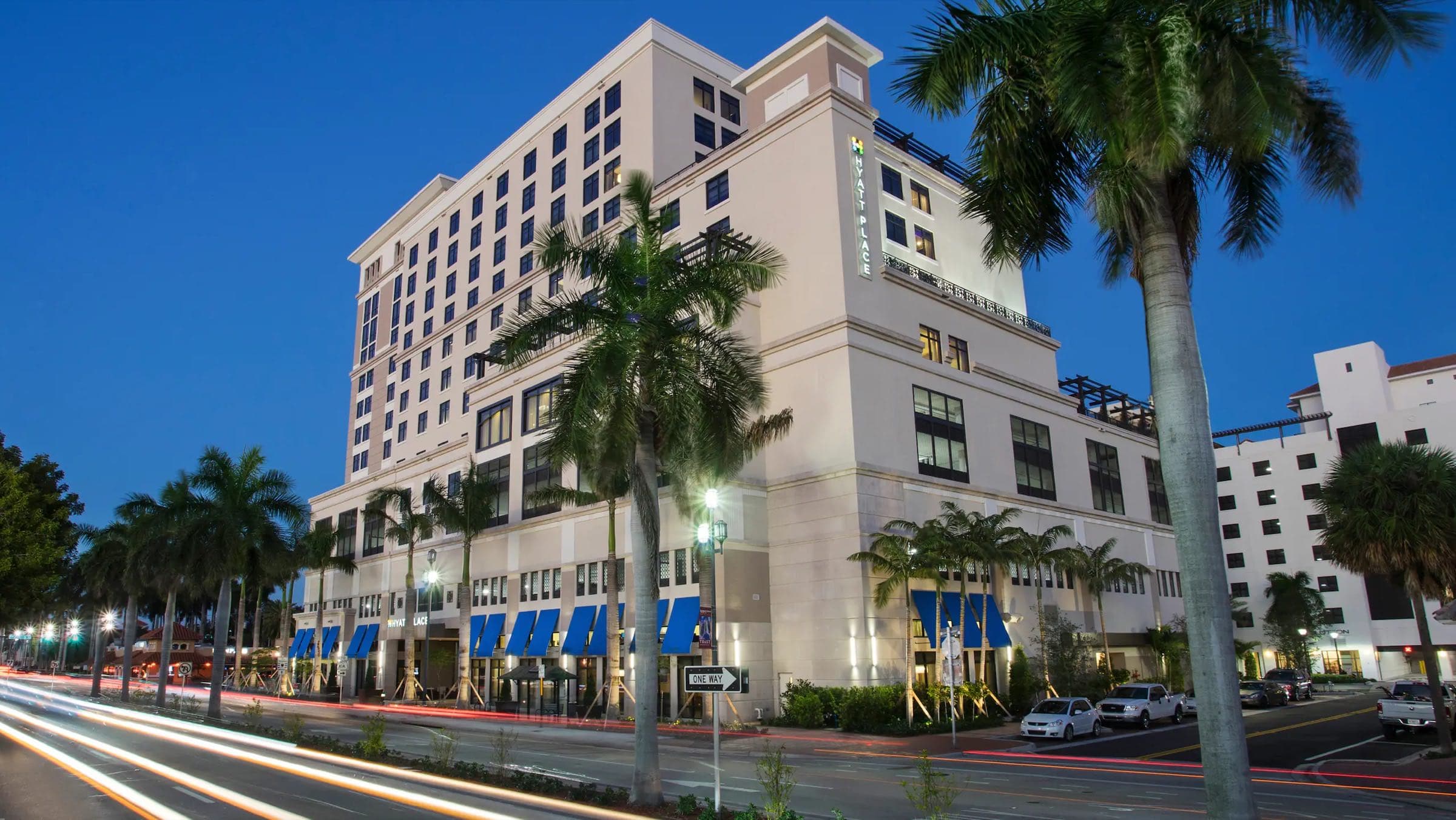 The Hyatt Place hotel building lit at night on a downtown Boca Raton corner, with royal palms lining the street frontage, blue storefront awnings, and light trails from passing traffic
