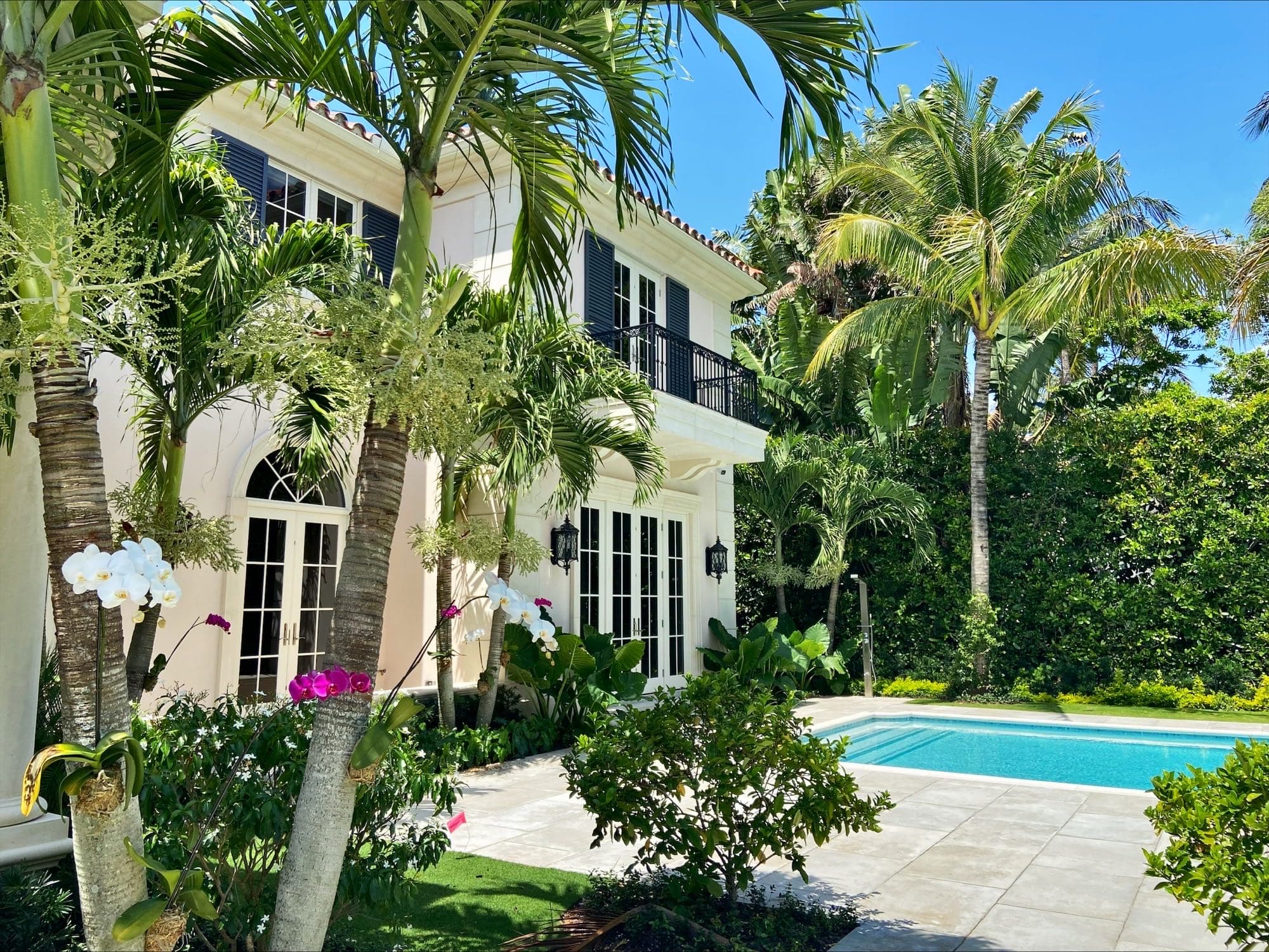 Two-story cream stucco residence with a red tile roof and iron balconies seen across a swimming pool framed by coconut palms and banana foliage